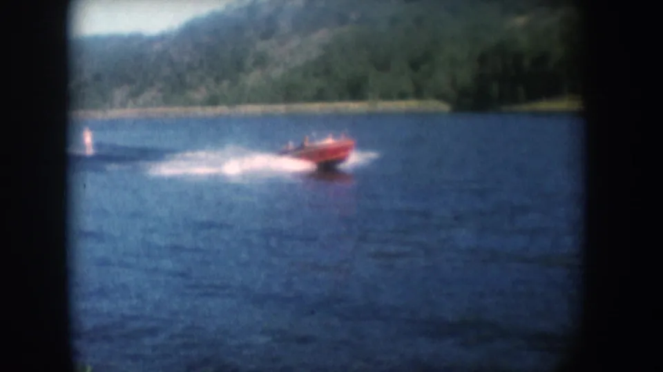 Red motorboat pulling a water skier across a South Dakota lake in 1956, with a wooded hillside beyond, shot on 8mm color film.