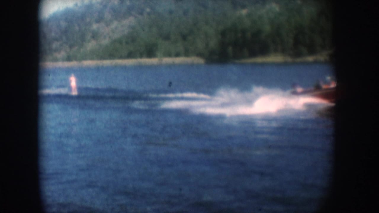 Water skier upright on lake surface with white spray; red motorboat at right edge of frame; wooded hills in background.