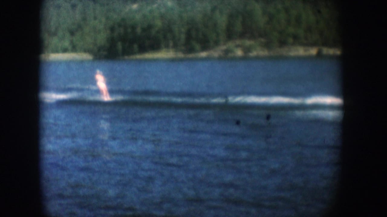 Water skier gliding across a South Dakota lake, circa 1956, with a low forested shoreline visible behind; shot on 8mm color film.