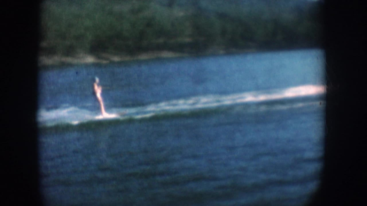 Water skier continuing across a South Dakota lake, circa 1956, spray wake visible, forested bank in background; 8mm color film.