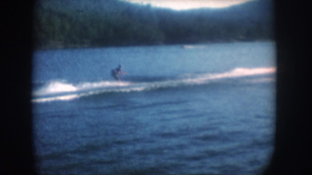 Distant water skier crossing a blue-tinted lake with a white wake, forested hillside and hills behind, 8mm film