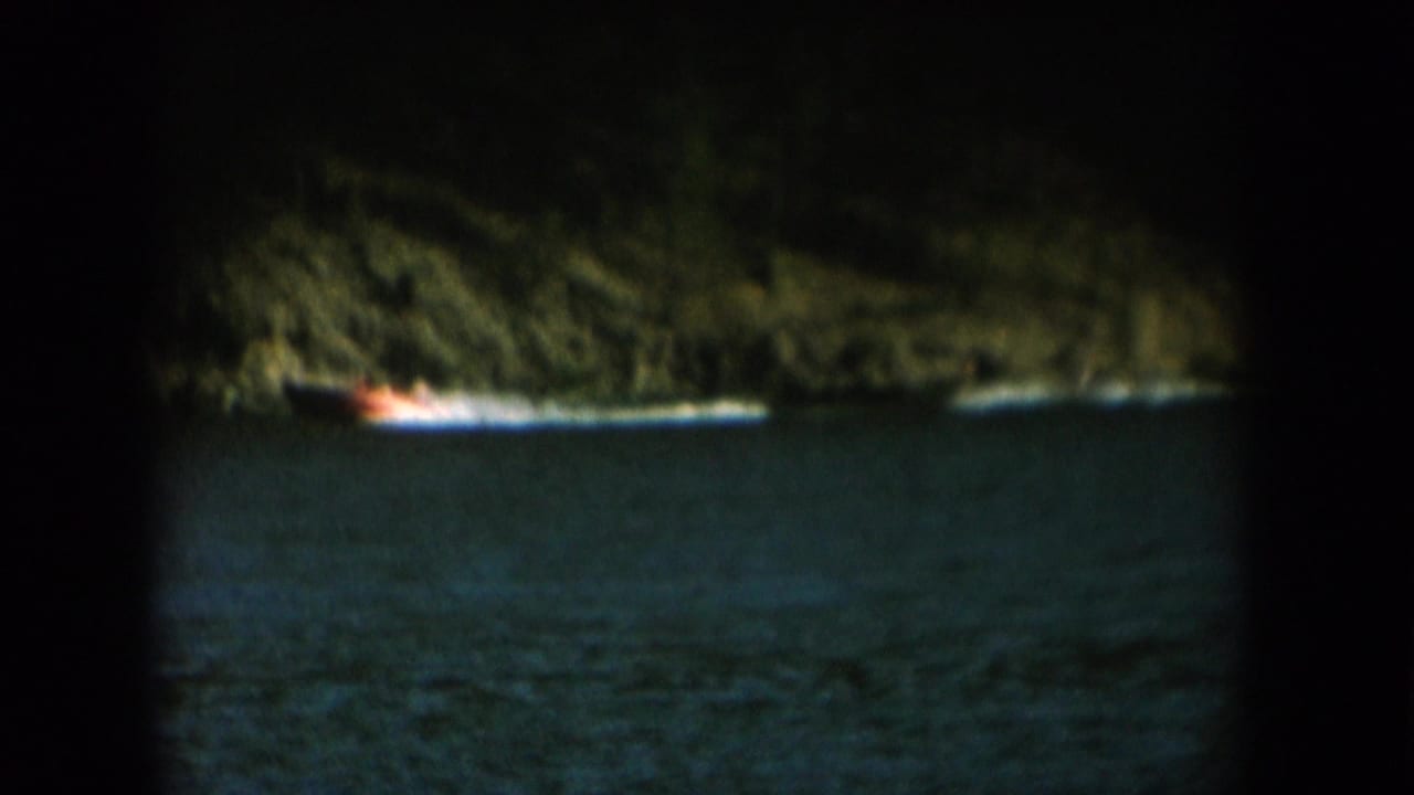 Low-light 8mm footage, likely dusk: steep wooded cliff above dark lake water with an orange-white boat light at the waterline, c. 1956