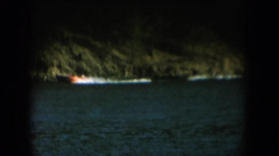 Low-light 8mm footage, likely dusk: steep wooded cliff above dark lake water with an orange-white boat light at the waterline, c. 1956