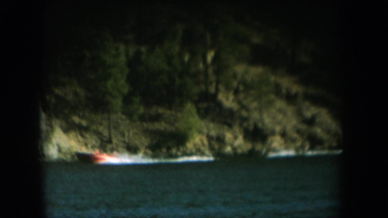 8mm home movie frame circa 1956 showing a red-orange speedboat cutting across a dark lake with a tree-covered cliff behind, deep circular vignette.