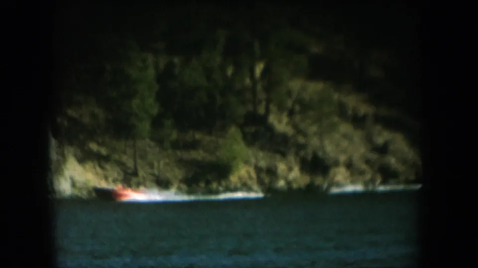 8mm home movie frame circa 1956 showing a red-orange speedboat cutting across a dark lake with a tree-covered cliff behind, deep circular vignette.