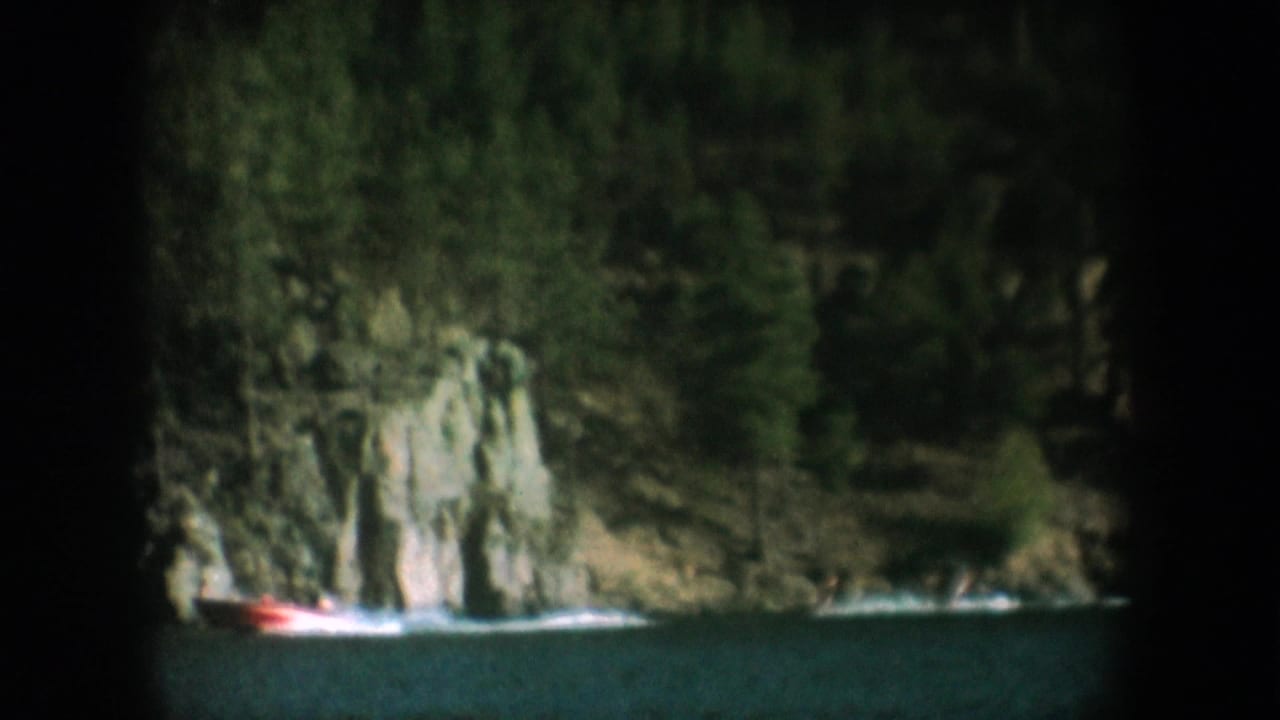 Mid-1950s 8mm home movie frame showing a speedboat passing a prominent exposed rock cliff on a dark lake, likely South Dakota, with characteristic vignetting.