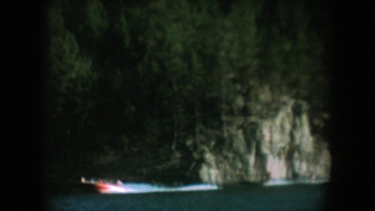 8mm home movie frame circa 1956 showing a red-orange speedboat in the lower frame on a dark lake with a rugged cliff and conifer forest, likely South Dakota.