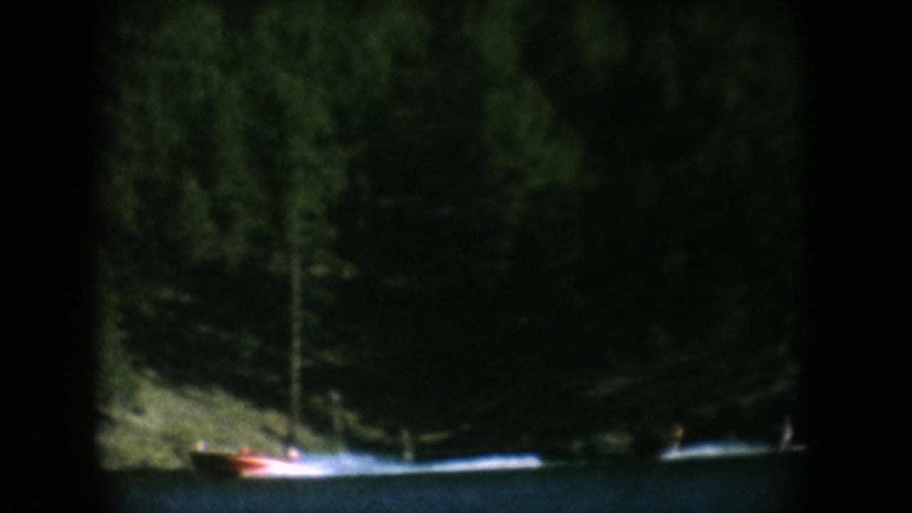 8mm home movie frame circa 1956 showing a distant speedboat on a dark lake with a prominent rocky spire and dense conifer forest backdrop.