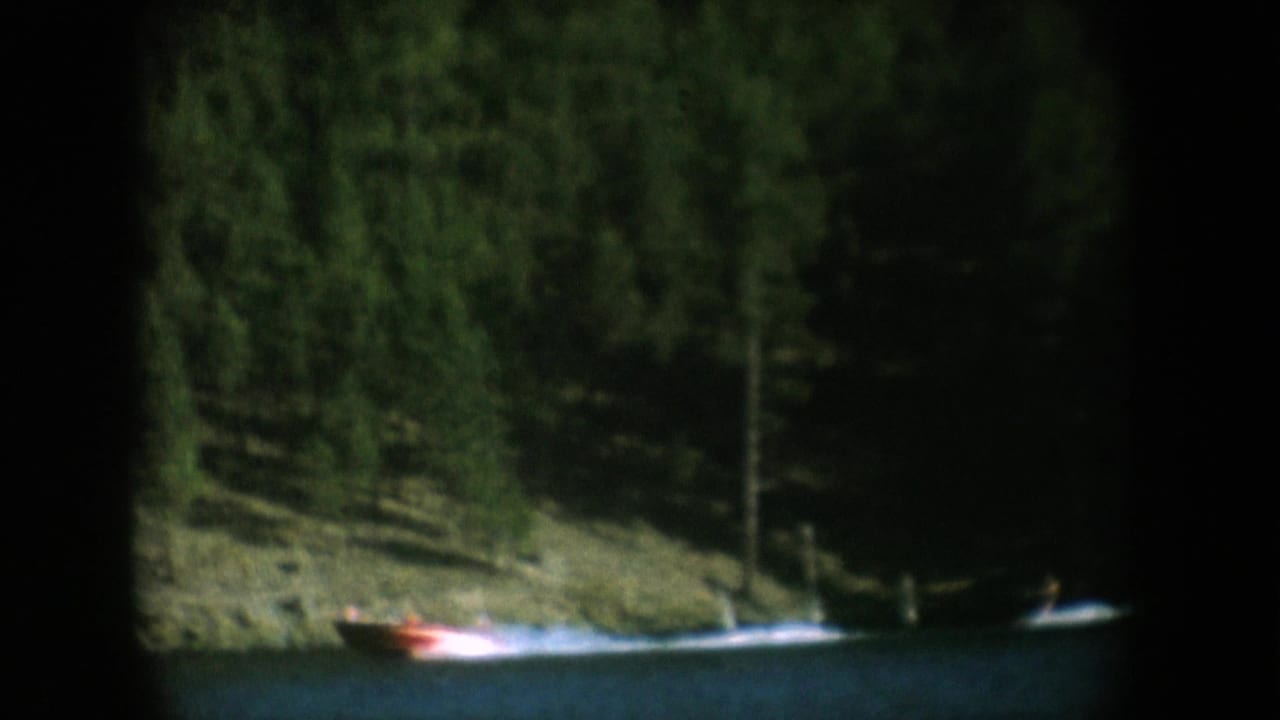 Red motorboat with white wake on a calm lake, coniferous forest shoreline behind, 8mm home movie footage from what may be South Dakota circa 1956.