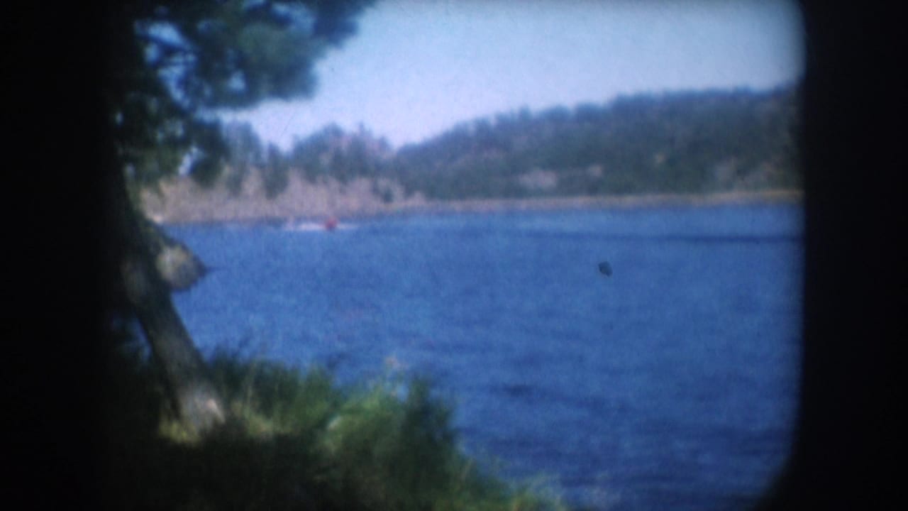 Wide lake view with rocky forested hillside across the water, small red boat in mid-distance, pine branches at right foreground, dark vignette framing.