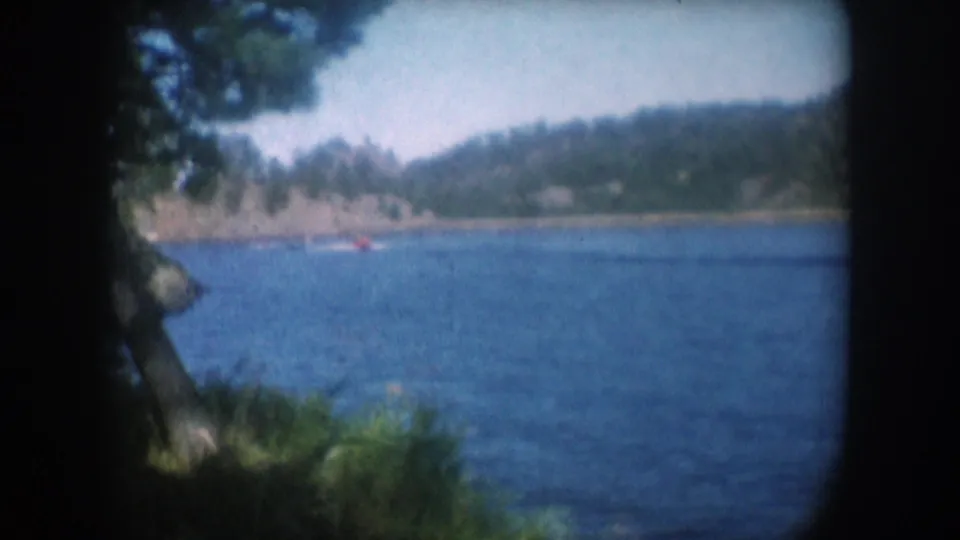 Lake scene viewed through a pine-shaded shore position with rocky hills across the water and a small motorboat visible, 8mm home movie circa 1956.
