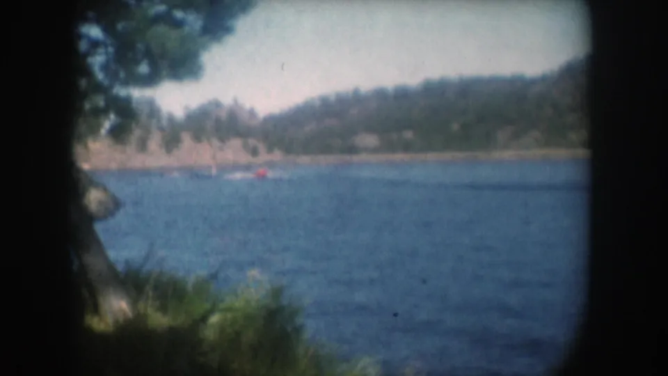 Final wide lake view with the boat receding into the distance, pine tree foreground framing, rocky wooded slopes across the water, 8mm home movie circa 1956.