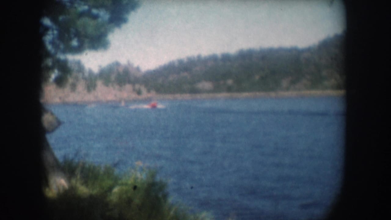 Red motorboat crossing a lake, possibly South Dakota circa 1956, viewed through 8mm film-gate borders with green foreground foliage at lower left.
