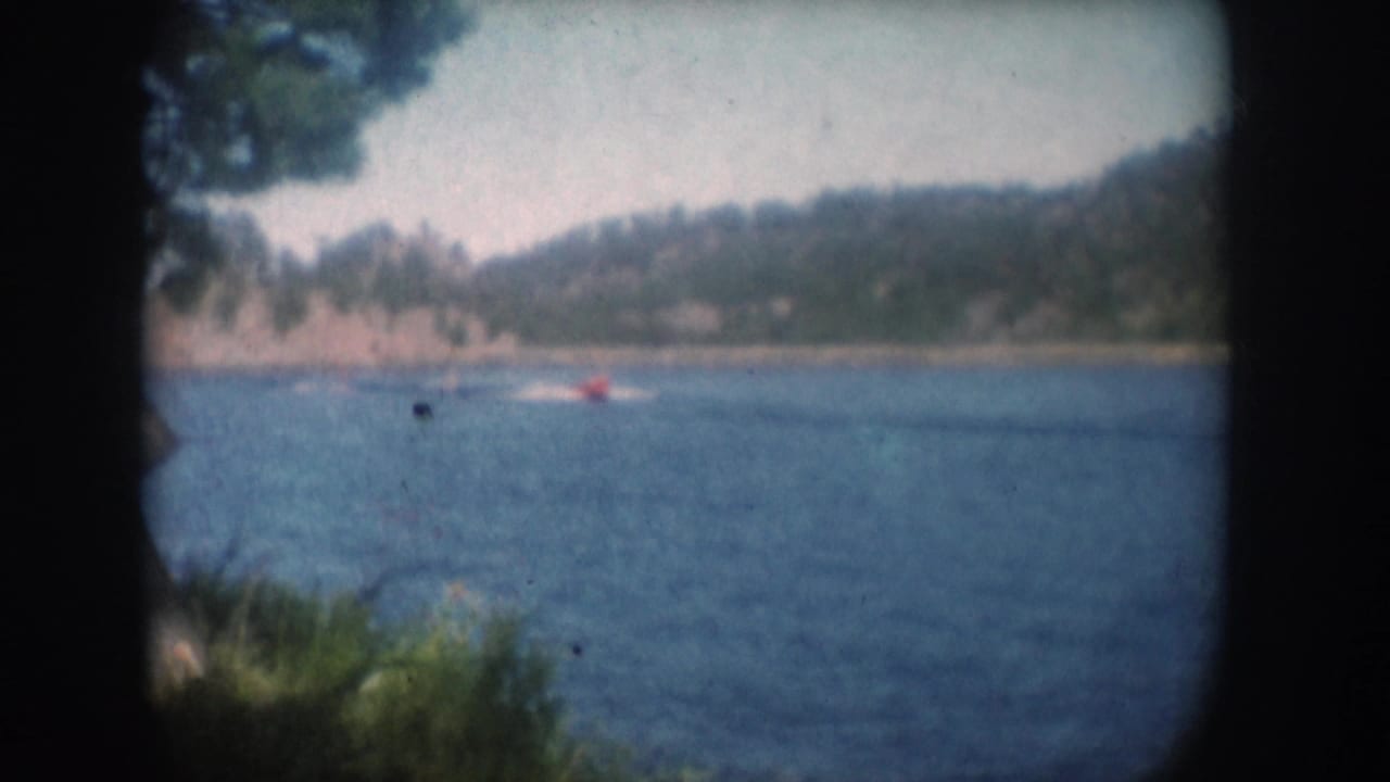 Red motorboat in mid-distance on a lake, film-gate borders visible, right-side foliage and wooded shoreline in background.