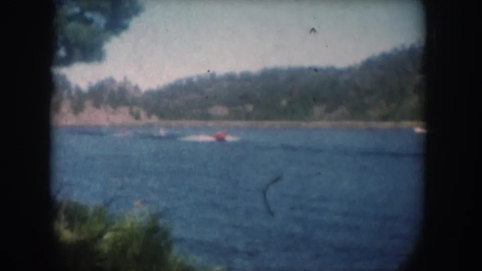 Red motorboat tracked across open water, possibly South Dakota circa 1956, with green foreground vegetation and a forested hillside shoreline.