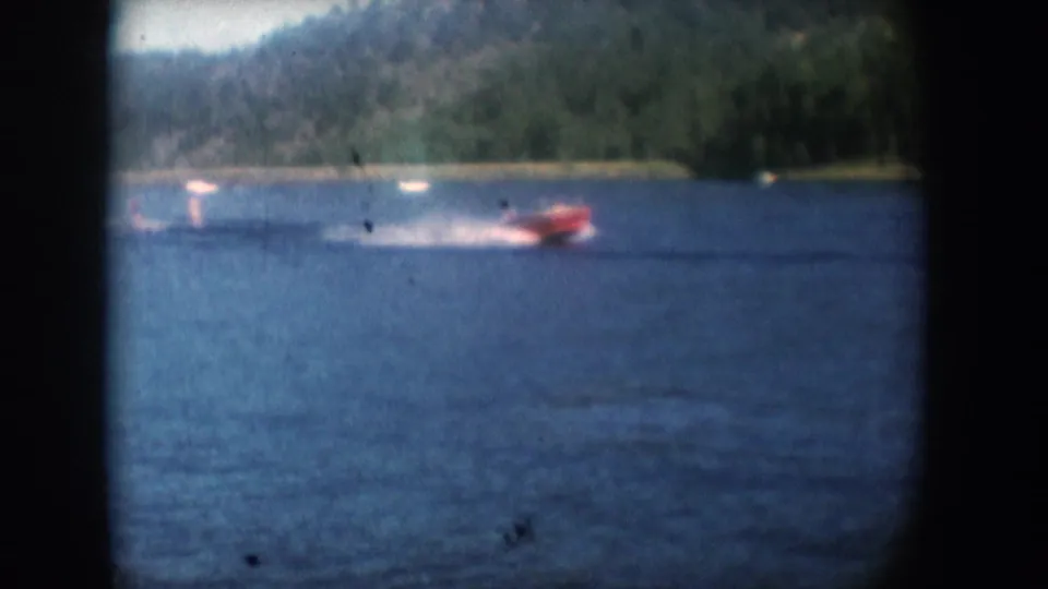 Red motorboat with prominent white wake on a lake, circa mid-1950s 8mm footage; boat is close enough that occupants may be present, though not identifiable.
