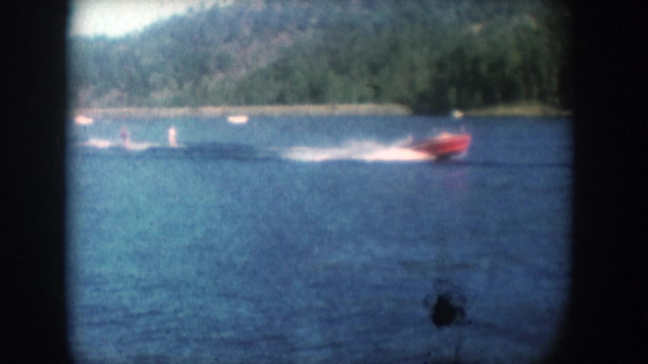 1956 8mm home movie frame: red motorboat towing water skiers on a South Dakota lake, tree-lined far shore, film grain and emulsion spot visible.