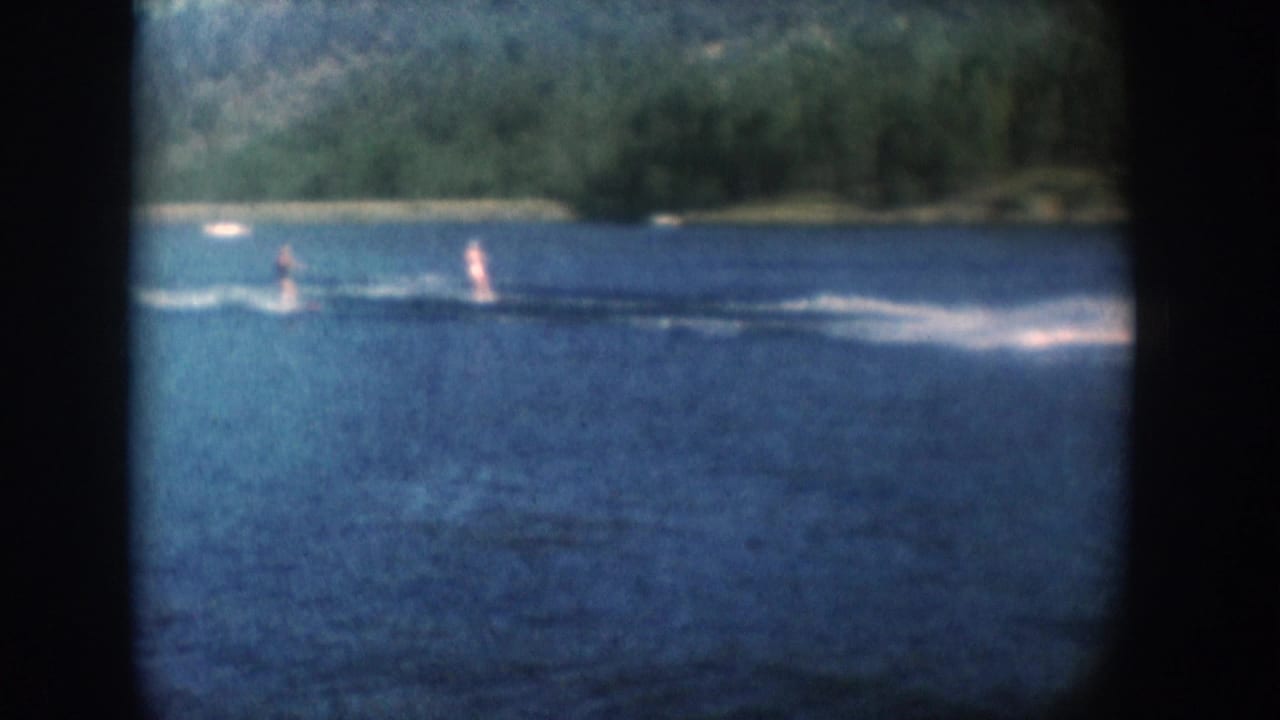 Two water skiers leaving white wakes across a blue lake, forested shoreline in background, 8mm vignette border visible.