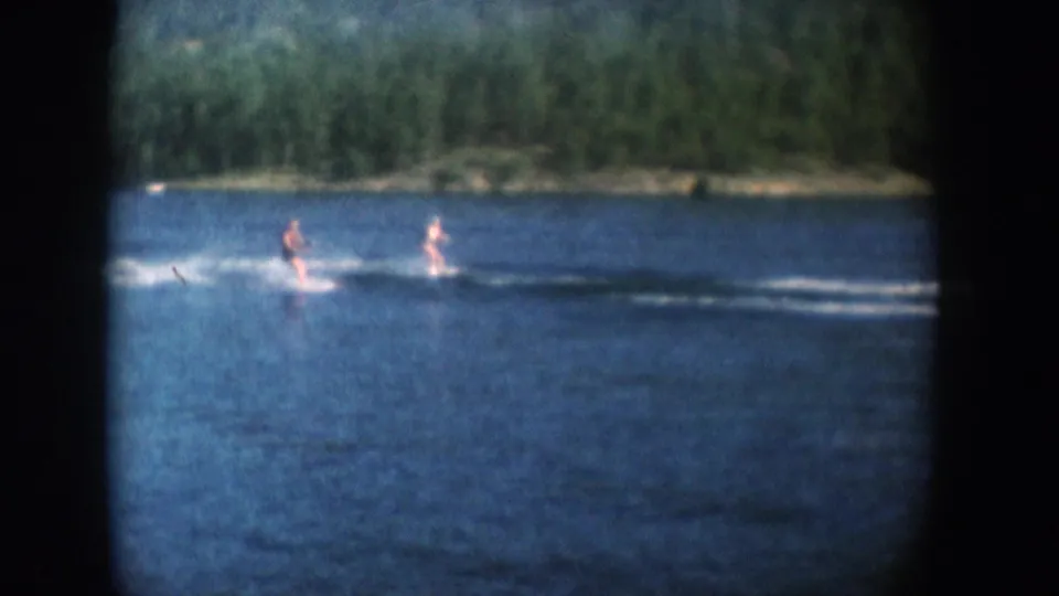 1956 8mm home movie frame: water skier on a South Dakota lake with dense green reeds and trees along the near shore.