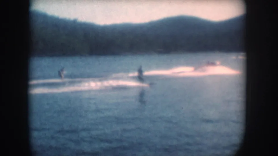 1956 8mm home movie frame: water skier on a South Dakota lake below low wooded hills, pale sky, film grain and vignette.