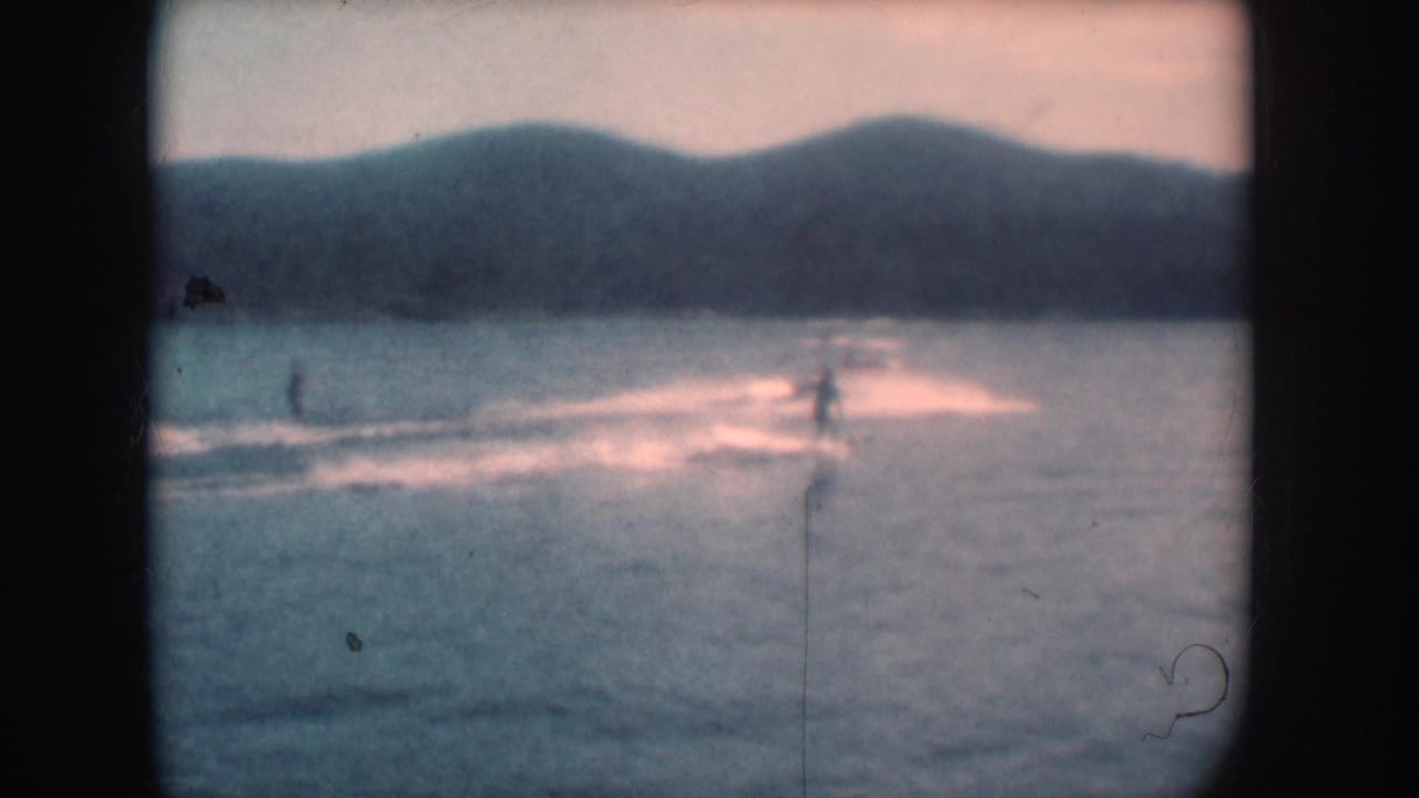 8mm home movie frame circa 1956 showing a water skier in the middle distance on a South Dakota lake, low hills across the far shore, warm faded emulsion