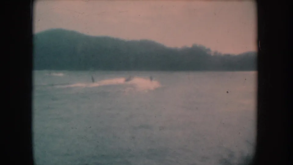 8mm home movie frame circa 1956 showing a lake and gently rolling far-shore hills; a small figure may be visible on the water surface amid heavy grain