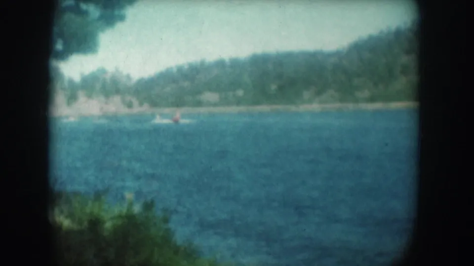 8mm home movie frame circa 1956 showing a teal-blue lake with tree-covered hills, leafy foreground growth, and a small red-orange object on the water surface