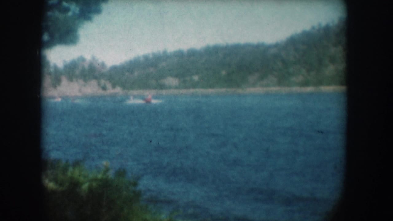 Wide 8mm shot from shore of a lone water skier in red on a lake in South Dakota, circa 1956, with wooded hillside and pale hazy sky.