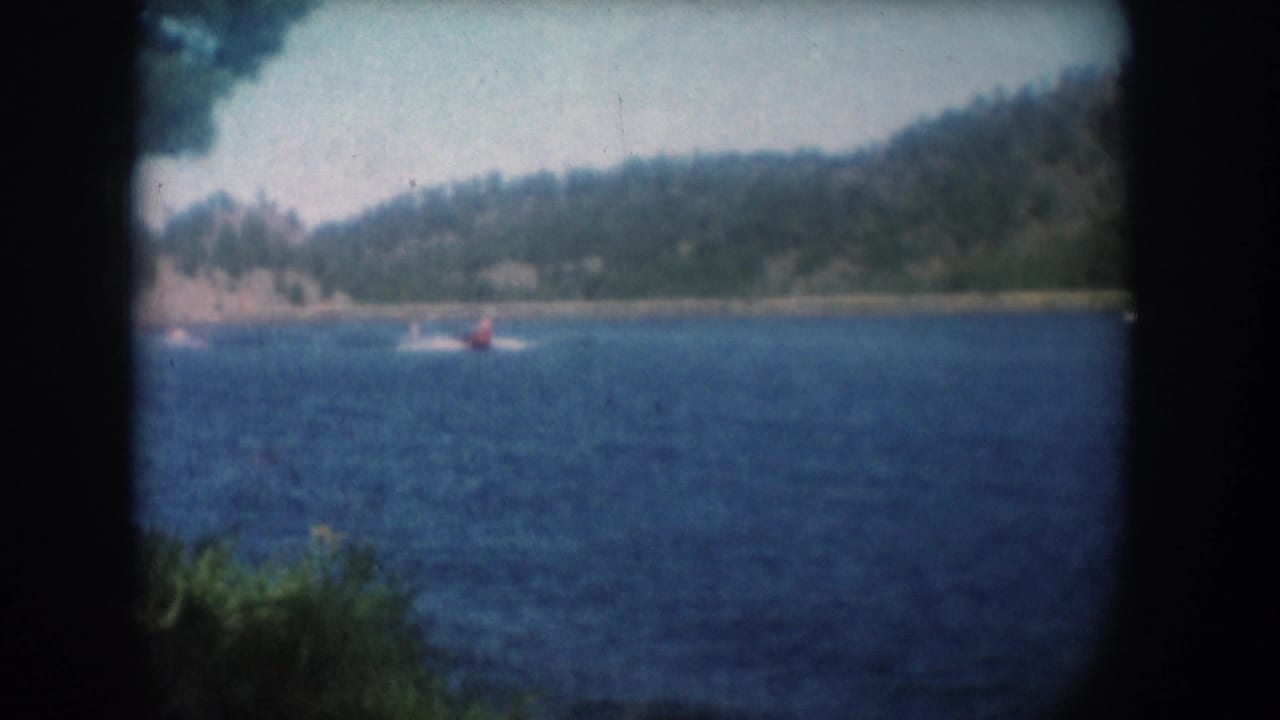 Same lake panorama; distant water skier in red has shifted position, tree-lined hills and pale sky visible through 8mm vignette.