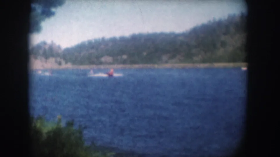 8mm color film frame of a water skier in red on a South Dakota lake, circa 1956; rocky wooded shoreline and pale sky visible.