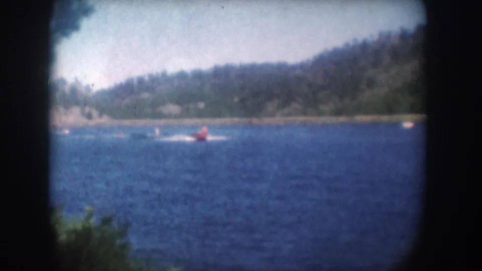 Closing 8mm frame of a water skier in red on a South Dakota lake, circa 1956; sky brightens at right above a forested hillside.
