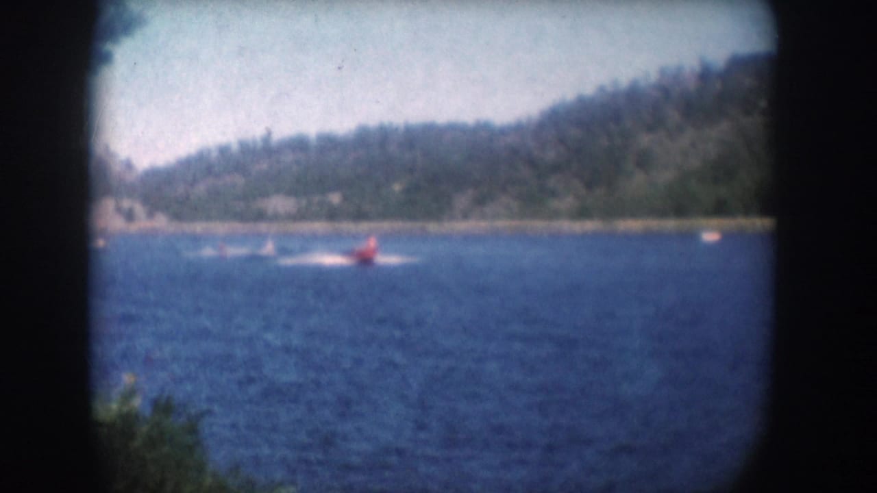 Red speedboat on a blue lake consistent with 1950s South Dakota recreational footage; dense forested hillside beyond; 8mm color film.