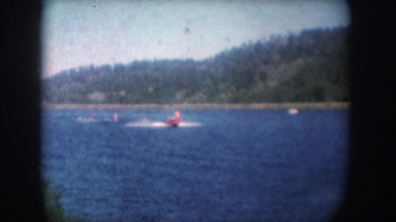 Red speedboat on blue lake with forested hill along far shore; 8mm grain and soft color fading visible.
