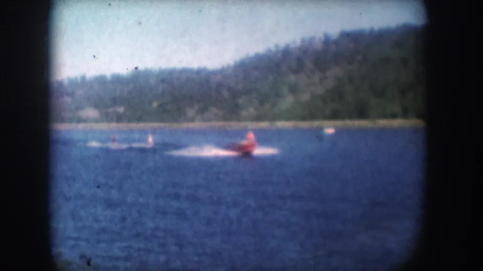 Red speedboat towing apparent water-skiers on a blue lake consistent with mid-1950s South Dakota; wooded hillside in background; 8mm film.