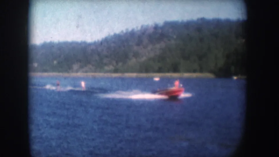 Red speedboat and trailing water-skiers on a blue lake consistent with 1950s South Dakota footage; dense forested hillside; 8mm color amateur film.