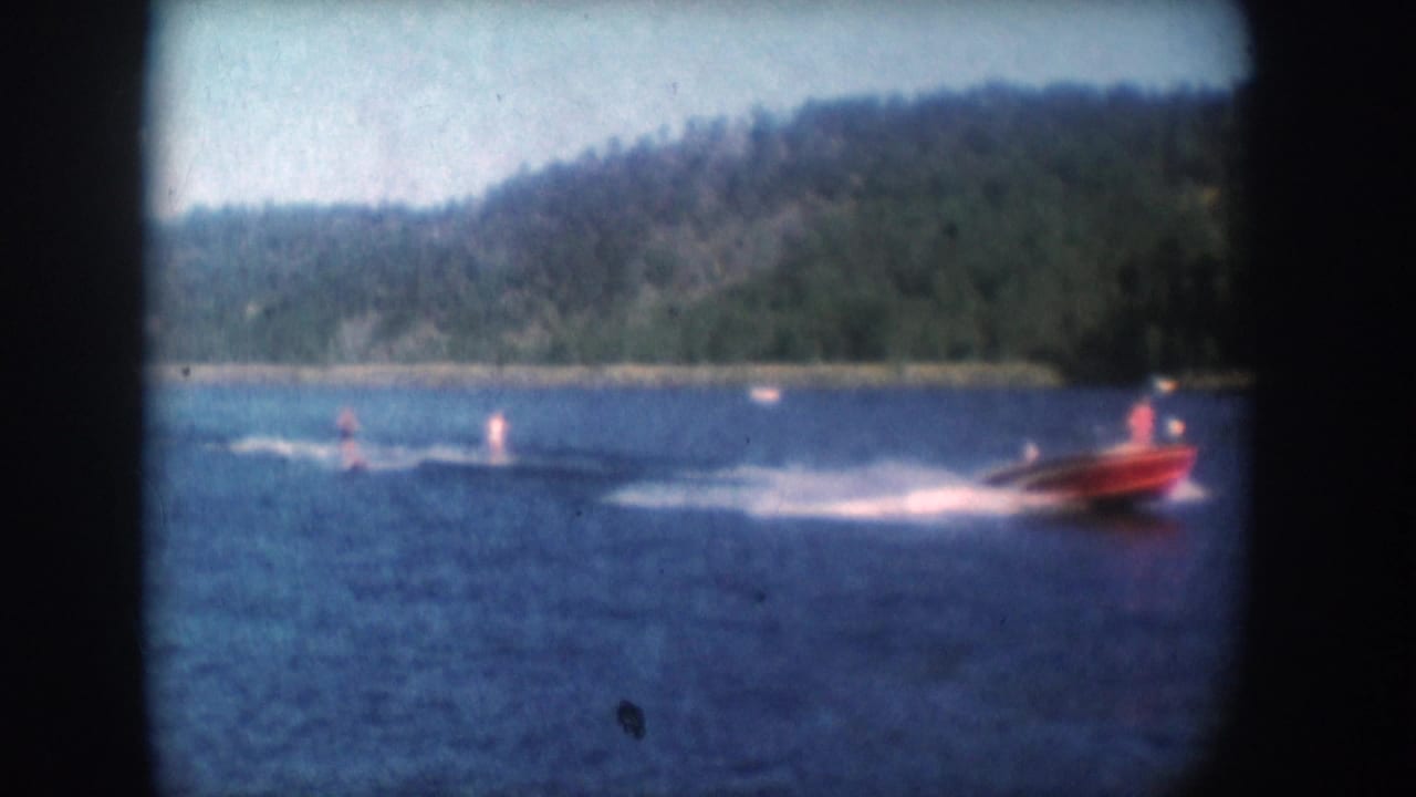Red motorboat towing water skiers on a lake described as South Dakota, USA, circa 1956; white wake visible and forested shore behind.