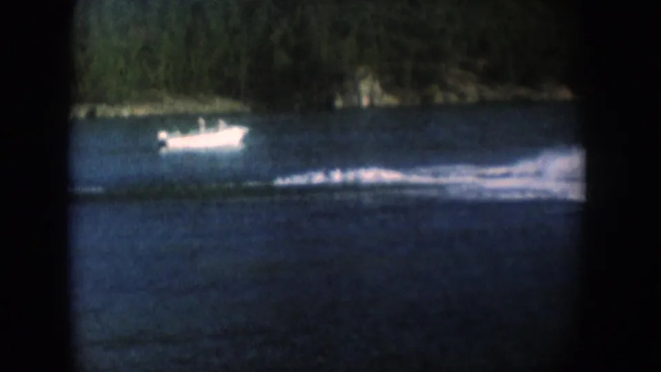 A pale motorboat on a lake with a forested shore, possibly South Dakota circa 1956; notable color desaturation and 8mm vignetting throughout.