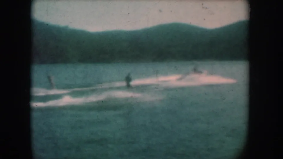 Two water skiers on an open lake described as South Dakota circa 1956; strong teal color shift from 8mm film aging, with rolling hills in the background.