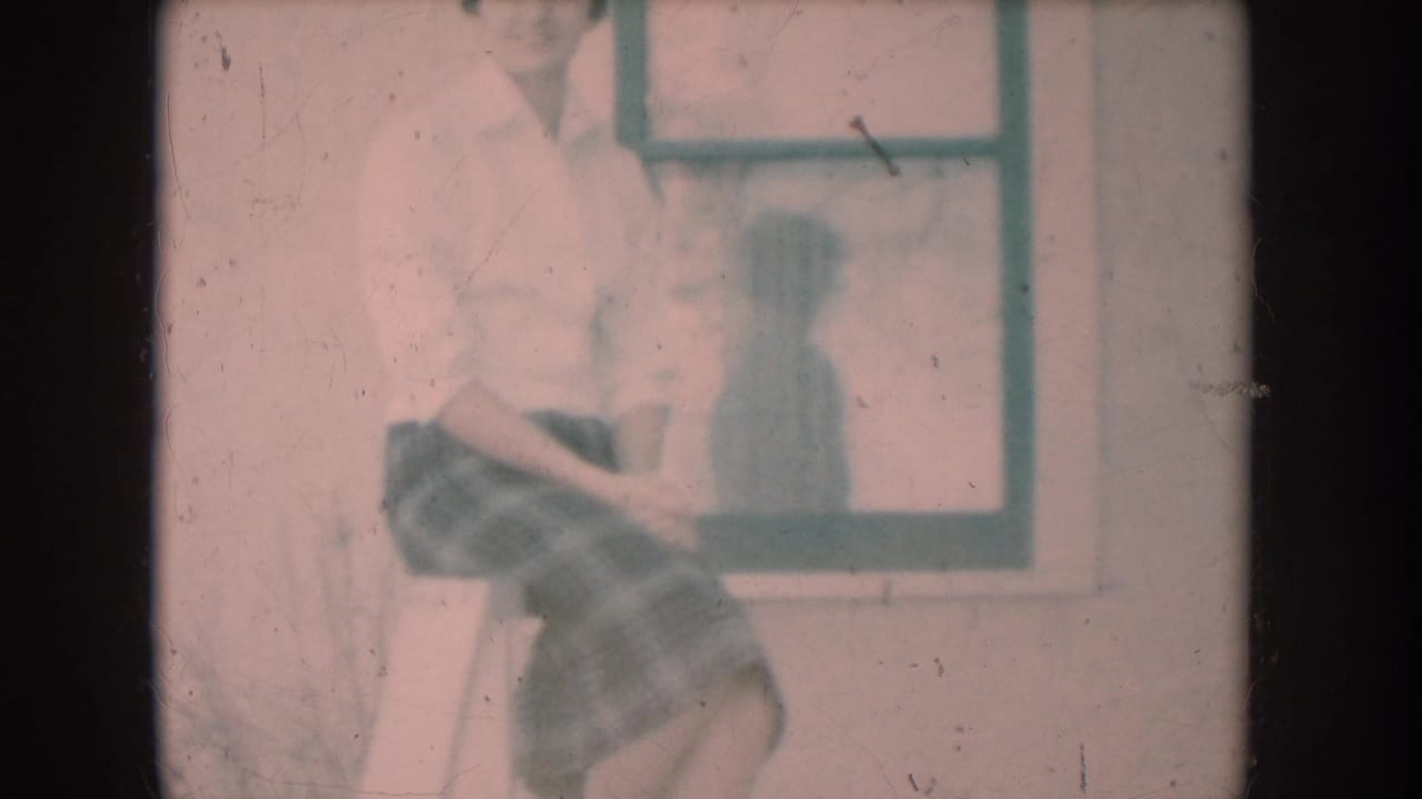 8mm home-movie frame c.1956: woman in white shirt and plaid skirt seated on porch railing, window with her reflection behind her, pink faded color cast.