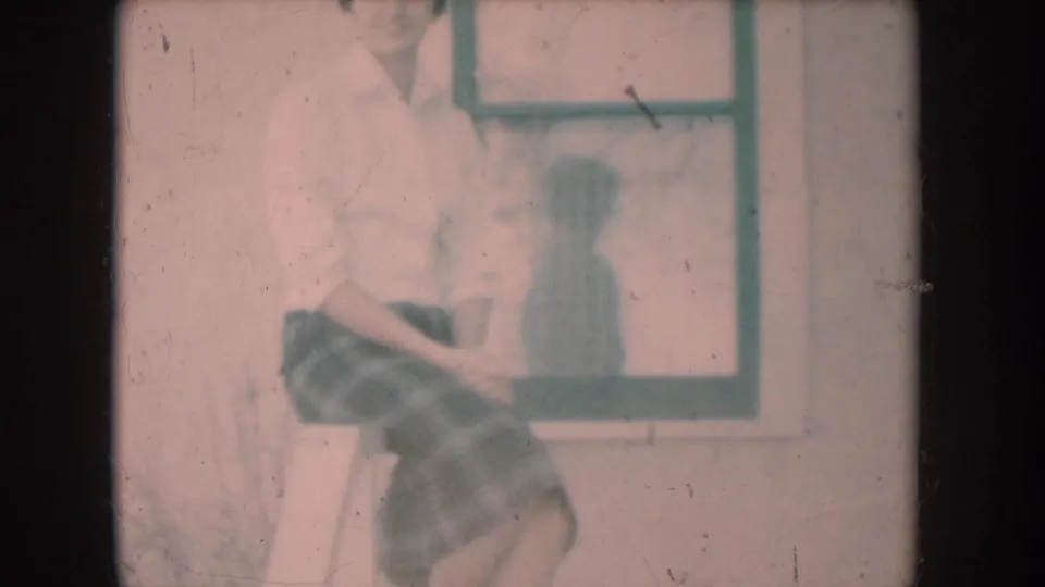 8mm home-movie frame c.1956: woman in white shirt and plaid skirt seated on porch railing, window with her reflection behind her, pink faded color cast.