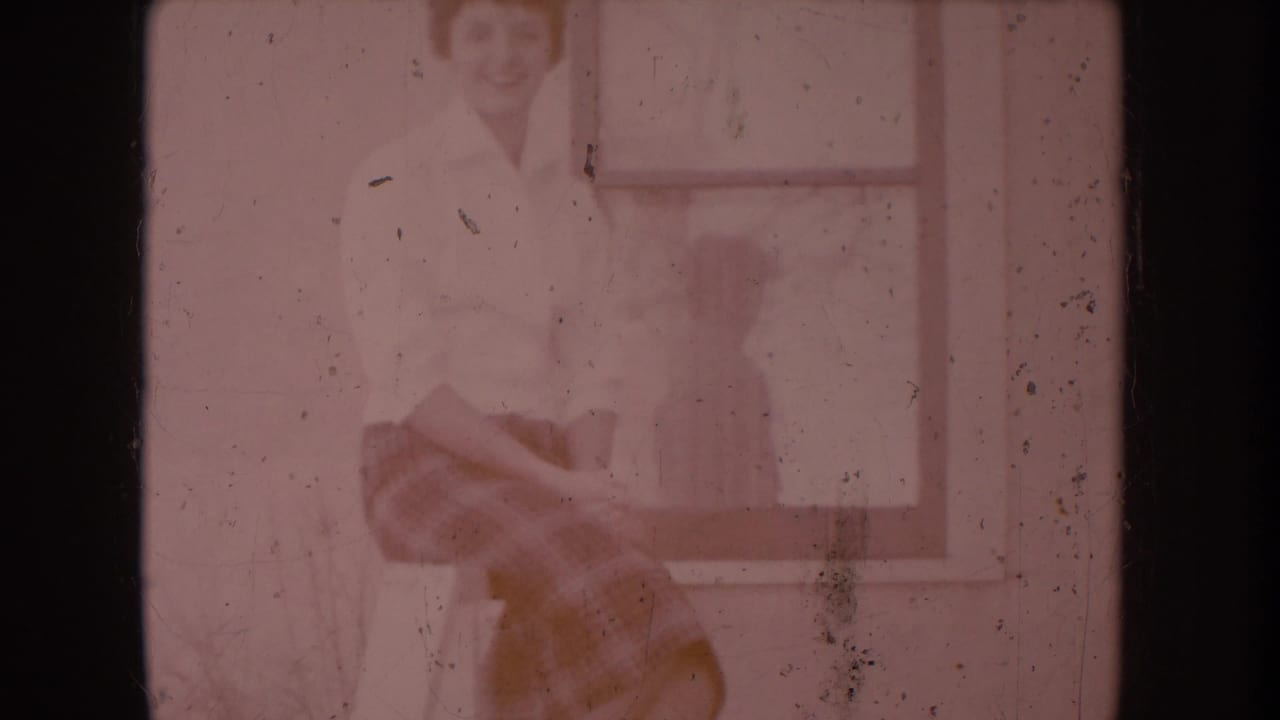 Woman in white shirt and plaid skirt on porch railing, stronger red-pink color cast, dust and grain, window with reflection behind her.