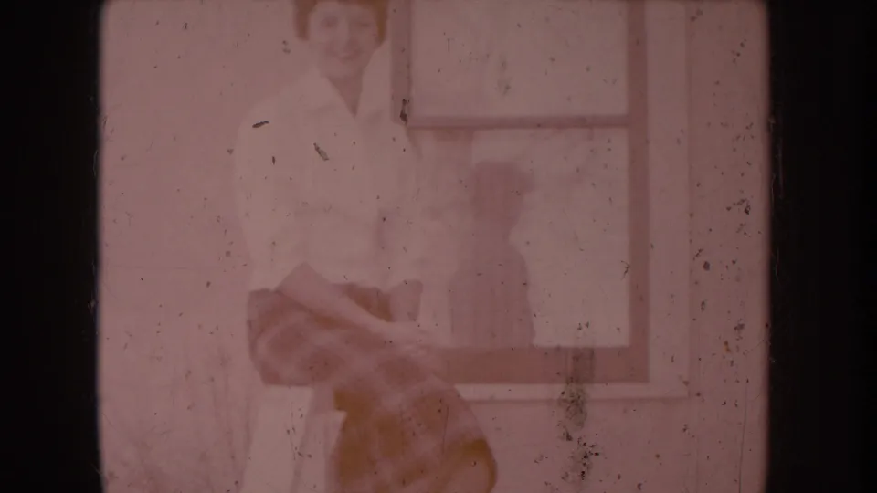 8mm home-movie frame c.1956: woman in white shirt on porch railing, pronounced red color shift with warm orange at lower edge, window reflection behind her.