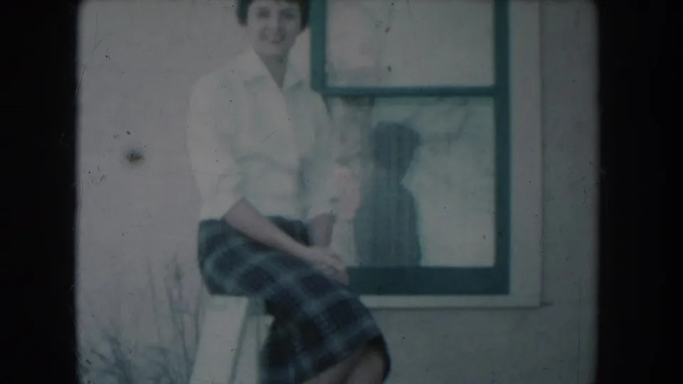 8mm home-movie frame c.1956: smiling woman in white shirt and plaid skirt on porch railing, blue-cyan color cast, window and her silhouette reflection visible behind her.
