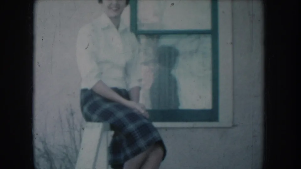 8mm home-movie frame c.1956: woman smiling on porch railing in white shirt and plaid skirt, teal-painted window frame and her reflection visible behind her, blue color cast.