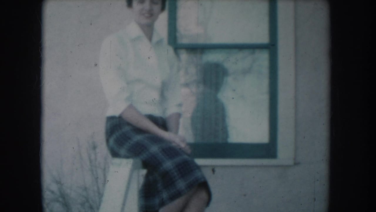 A woman in 1950s casual dress, white button-up shirt and plaid skirt, sits on a porch railing beside a white-framed window, in what appears to be a South Dakota residential setting circa 1956.