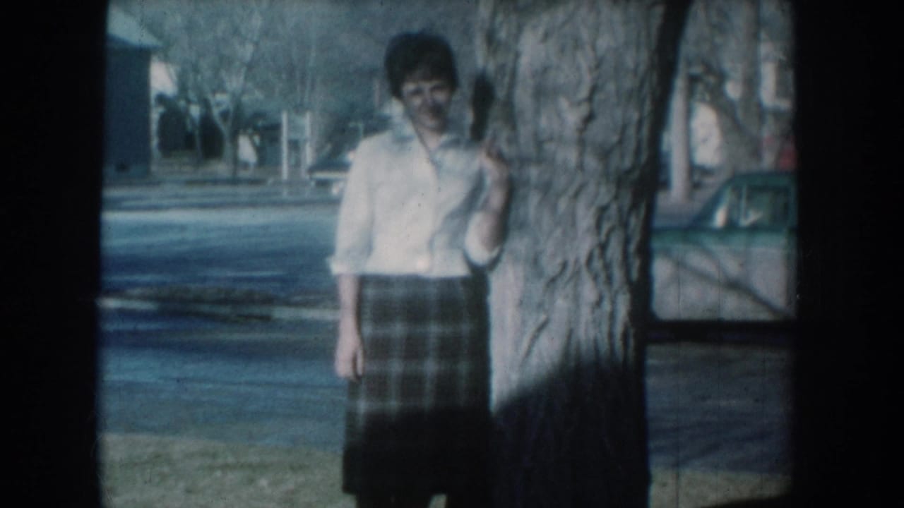 Woman in white shirt and plaid skirt standing beside a large tree trunk on a residential street.