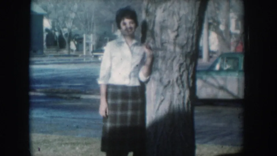 A smiling woman in a white shirt and plaid skirt stands beside a large tree; a residential street with period vehicles is visible behind her, consistent with mid-1950s South Dakota.