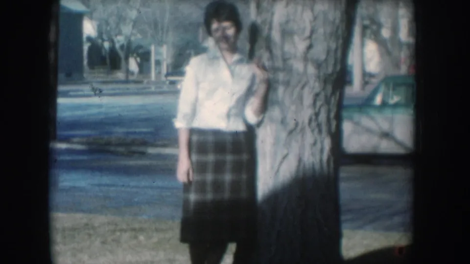 A woman in a white collared shirt and plaid skirt poses beside a large mature tree on a quiet residential street; parked cars and houses typical of a mid-1950s American town are visible behind her.