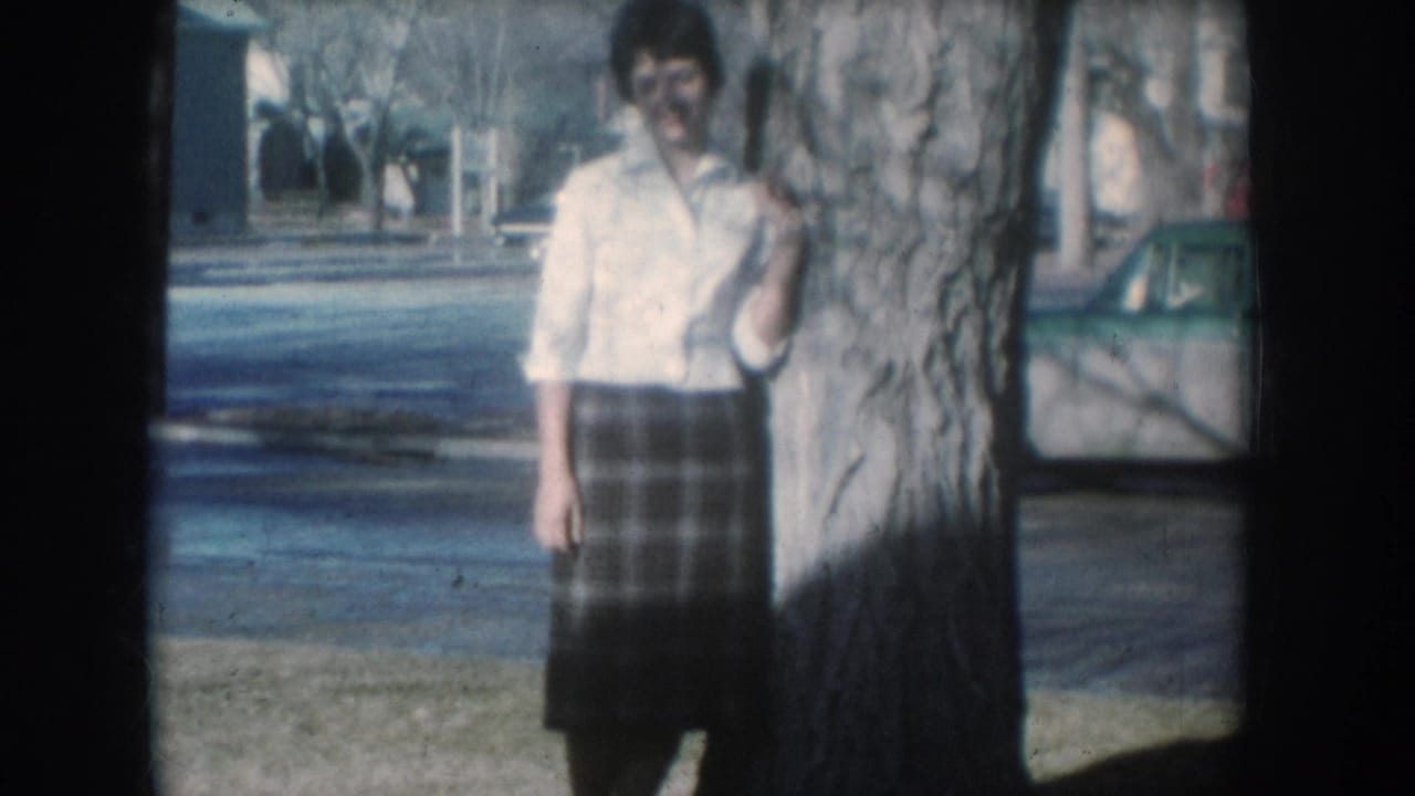 Young woman in white shirt and plaid skirt poses beside a large tree on a quiet 1950s suburban American street, mid-century cars faintly visible in background.