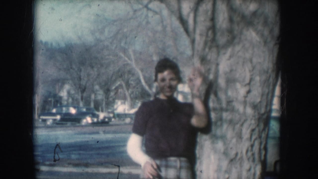 Woman in a dark short-sleeved top and plaid skirt waves at the camera while leaning against a tree; rounded mid-century cars and bare trees are visible on the residential street behind her.